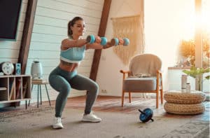 athletic girl in leggings and top crouches with dumbbells at home.